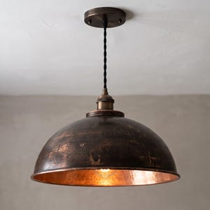 Puede incluir: Una lámpara colgante en forma de cúpula con un acabado exterior de bronce oscuro desgastado y un interior de cobre pulido cálido. La lámpara cuelga de un cable negro trenzado unido a un soporte de techo circular de bronce a juego.