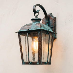 May include: A wall-mounted lantern with a weathered copper finish featuring green patina accents. The fixture has a curved arm, a rectangular frame with clear textured glass panels, and an exposed Edison-style light bulb glowing warmly inside.