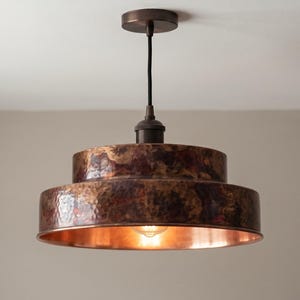 May include: A tiered pendant light fixture featuring a dark, hammered copper finish on the exterior and a smooth, reflective copper interior. The lamp is suspended by a black cord from a matching dark copper circular ceiling canopy mount.