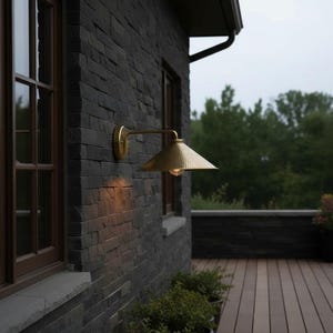 May include: Exterior wall with a brass-colored, cone-shaped outdoor light fixture. The wall is made of dark gray stone bricks. A wooden-framed window is visible to the left. The background features a wooden deck and lush green trees.