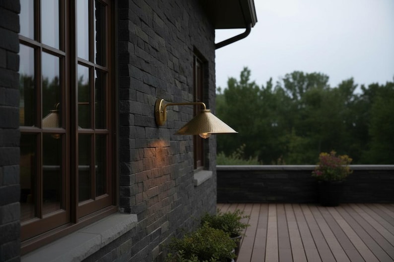 May include: Exterior wall with a brass-colored, cone-shaped outdoor light fixture. The wall is made of dark gray stone bricks. A wooden-framed window is visible to the left. The background features a wooden deck and lush green trees.