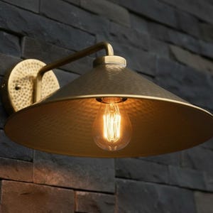 May include: A brass-colored wall sconce with a conical shade and a decorative, textured surface. The sconce features a vintage-style lightbulb and is mounted on a dark, textured stone wall. The fixture has a curved arm.