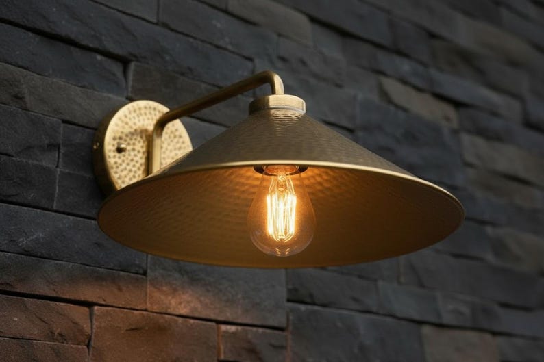 May include: A brass-colored wall sconce with a conical shade and a decorative, textured surface. The sconce features a vintage-style lightbulb and is mounted on a dark, textured stone wall. The fixture has a curved arm.