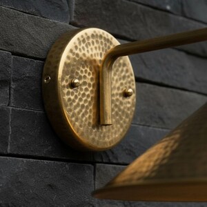 May include: A brass wall sconce with a hammered texture, mounted on a dark gray stone wall. The sconce features a circular backplate, a curved arm, and a conical shade. The light fixture is designed for indoor or outdoor use.