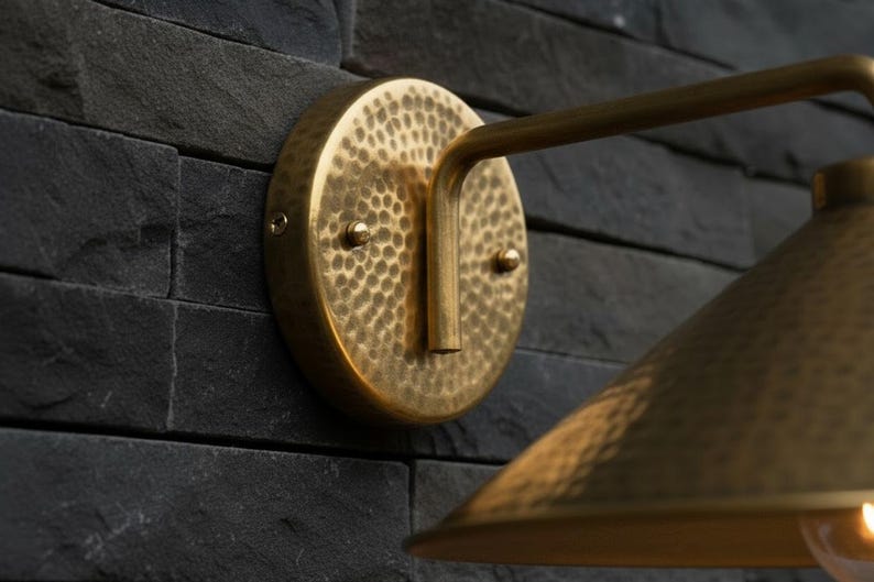 May include: A brass wall sconce with a hammered texture, mounted on a dark gray stone wall. The sconce features a circular backplate, a curved arm, and a conical shade. The light fixture is designed for indoor or outdoor use.