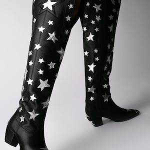 May include: Black knee-high boots featuring a textured leather finish and numerous silver star appliques scattered across the shaft and foot. The boots have a pointed toe with a silver metal cap, a block heel, and a tall, curved top edge.