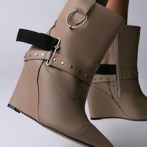 Peut inclure: Une paire de bottines compensées taupe avec un bout pointu, une fermeture éclair latérale et une bride décorative ornée de clous argentés. Les bottines présentent un détail de bande élastique noire et un anneau métallique argenté sur la tige.