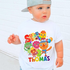 May include: A white short-sleeved t-shirt featuring a colorful graphic print of cartoon characters, balloons, and a rainbow. The text reads SUPER BDAY, the number 3, and the name THOMAS in vibrant, multi-colored letters across the front chest area.
