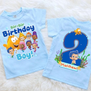May include: Two light blue toddler t-shirts. One shirt features a cartoon fish and characters with the words "bir-bir Birthday Boy!". The other shirt has a large blue number 2 with a cartoon character and the name "Matthew".