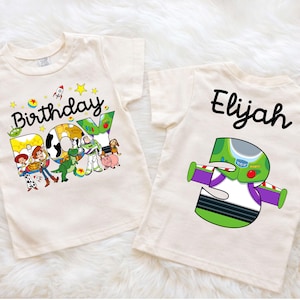 May include: Two cream-colored t-shirts. One features the text "Birthday BOY" with Toy Story characters. The other shirt has the name "Elijah" and a number "3" designed as Buzz Lightyear. Both shirts are short-sleeved.