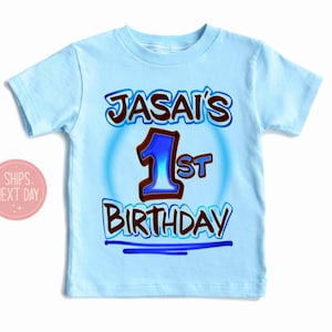 May include: Light blue t-shirt with the text "JASAI'S 1st BIRTHDAY" in brown and blue lettering. The number "1" is in blue with a brown outline. The shirt is a short-sleeved crew neck.