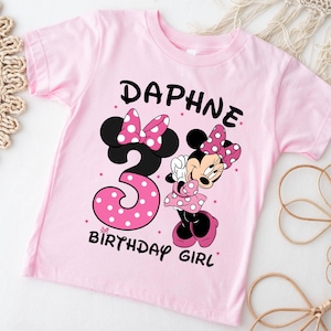 May include: A light pink t-shirt featuring a Minnie Mouse graphic. The shirt has the name "DAPHNE" above a large number "3" with pink polka dots, and the words "BIRTHDAY GIRL". Minnie Mouse is also featured on the shirt.