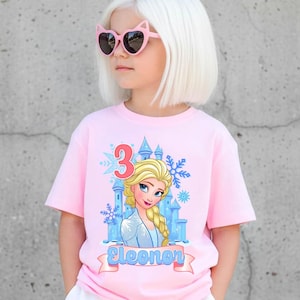Może przedstawiać: R&oacute;żowa koszulka z kresk&oacute;wkowym wizerunkiem księżniczki, niebieskim zamkiem, płatkami śniegu i cyfrą 3. Imię "Eleonor" jest wydrukowane na banerze. Dziecko ma na sobie r&oacute;żowe okulary przeciwsłoneczne w kształcie kocich oczu.