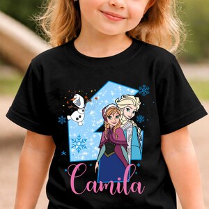 May include: A black short-sleeve t-shirt featuring a large light blue number four graphic with snowflakes, a cartoon snowman, and two animated characters. The name Camila is printed in pink cursive script at the bottom of the graphic design.