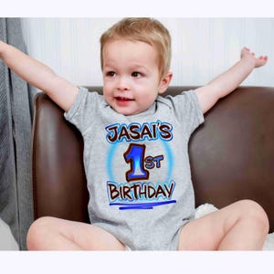 May include: A gray onesie with the text "JASAI'S 1st BIRTHDAY" in blue and brown lettering. The number 1 is in blue with a brown outline. The onesie is short-sleeved and the child is sitting on a brown chair.