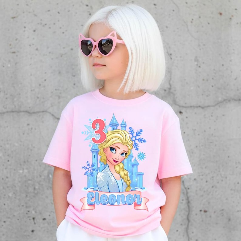 Może przedstawiać: R&oacute;żowa koszulka z kresk&oacute;wkowym wizerunkiem księżniczki, niebieskim zamkiem, płatkami śniegu i cyfrą 3. Imię "Eleonor" jest wydrukowane na banerze. Dziecko ma na sobie r&oacute;żowe okulary przeciwsłoneczne w kształcie kocich oczu.