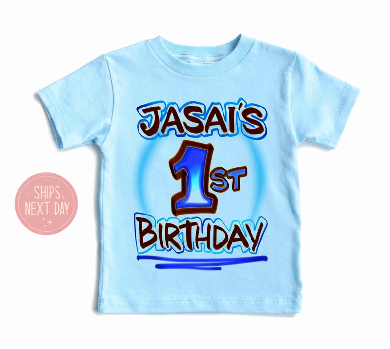 May include: Light blue t-shirt with the text "JASAI'S 1st BIRTHDAY" in brown and blue lettering. The number "1" is in blue with a brown outline. The shirt is a short-sleeved crew neck.
