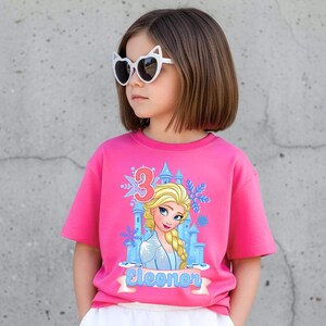 Może przedstawiać: Młoda dziewczyna w r&oacute;żowej koszulce z grafiką księżniczki z kresk&oacute;wki, zamkiem i cyfrą 3. Na koszulce widnieje r&oacute;wnież imię "Eleonor". Ma na sobie okulary przeciwsłoneczne w kształcie kocich oczu.