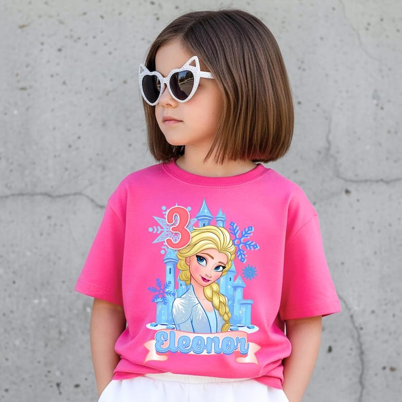 Może przedstawiać: Młoda dziewczyna w r&oacute;żowej koszulce z grafiką księżniczki z kresk&oacute;wki, zamkiem i cyfrą 3. Na koszulce widnieje r&oacute;wnież imię "Eleonor". Ma na sobie okulary przeciwsłoneczne w kształcie kocich oczu.