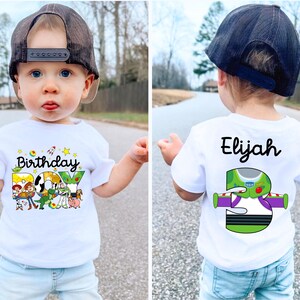 May include: A white t-shirt with a colorful "Birthday Boy" graphic featuring Toy Story characters. The back of the shirt has the name "Elijah" and a "3" with a Buzz Lightyear design. The child is wearing a black baseball cap.