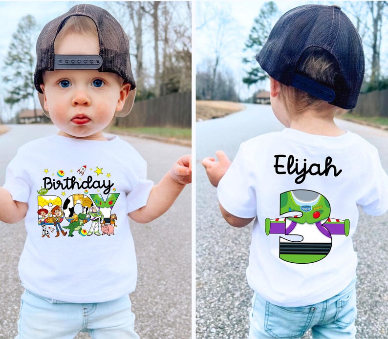 May include: A white t-shirt with a colorful "Birthday Boy" graphic featuring Toy Story characters. The back of the shirt has the name "Elijah" and a "3" with a Buzz Lightyear design. The child is wearing a black baseball cap.