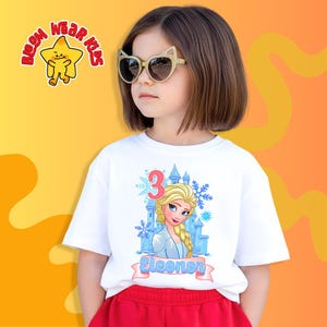 Może przedstawiać: Młoda dziewczyna w okularach przeciwsłonecznych w kształcie kocich oczu i białej koszulce z grafiką księżniczki z kresk&oacute;wki, cyfrą 3 i imieniem "Eleonor". Koszulka ma niebieski zamek i wz&oacute;r płatka śniegu. Ma na sobie czerwone szorty.