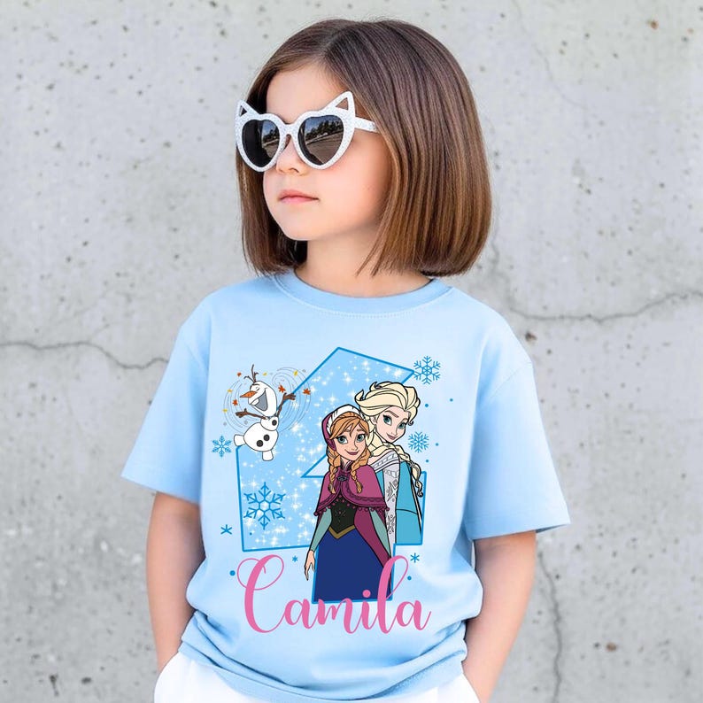 May include: A light blue short-sleeve t-shirt featuring a graphic print of the characters Anna, Elsa, and Olaf from Frozen. The design includes a large blue number four, snowflake accents, and the name Camila written in pink cursive script.
