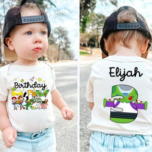 May include: A beige baby bodysuit with the words "Birthday BOY" and cartoon characters printed on the front. The back of the bodysuit has the name "Elijah" and a Buzz Lightyear graphic with the number "5". The child is wearing a black cap.