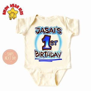 May include: A cream-colored baby onesie with the text "JASAI'S 1st BIRTHDAY" in blue and brown lettering. The number "1" is in blue with a blue outline. The onesie has short sleeves and a round neckline. The image also includes a cartoon star.