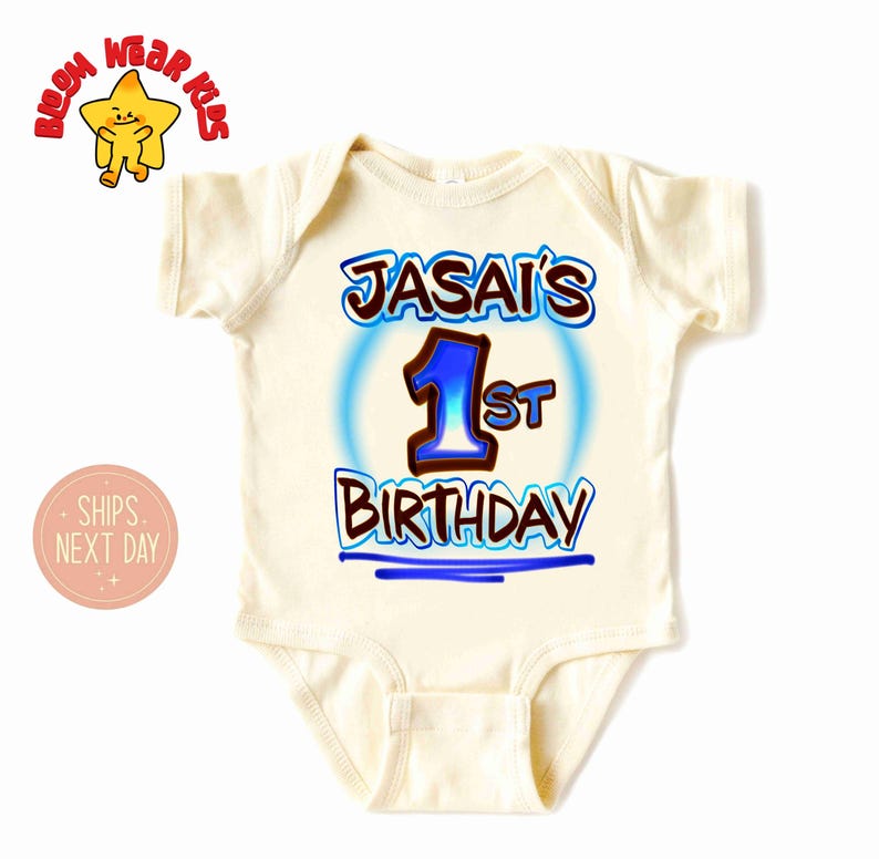 May include: A cream-colored baby onesie with the text "JASAI'S 1st BIRTHDAY" in blue and brown lettering. The number "1" is in blue with a blue outline. The onesie has short sleeves and a round neckline. The image also includes a cartoon star.