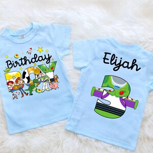 May include: Two light blue toddler t-shirts. One features the text "Birthday BOY" with Toy Story characters. The other shirt has the name "Elijah" and a Buzz Lightyear-themed number "3" graphic.