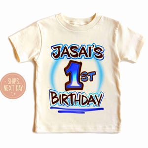 May include: Cream-colored short-sleeved t-shirt with the text "JASAI'S 1st BIRTHDAY" in blue and brown lettering. The number "1" is in blue with a brown outline, and the text is surrounded by a blue glow.