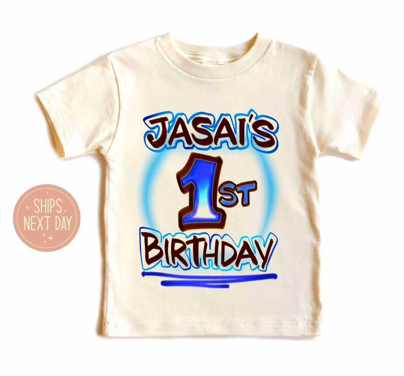 May include: Cream-colored short-sleeved t-shirt with the text "JASAI'S 1st BIRTHDAY" in blue and brown lettering. The number "1" is in blue with a brown outline, and the text is surrounded by a blue glow.