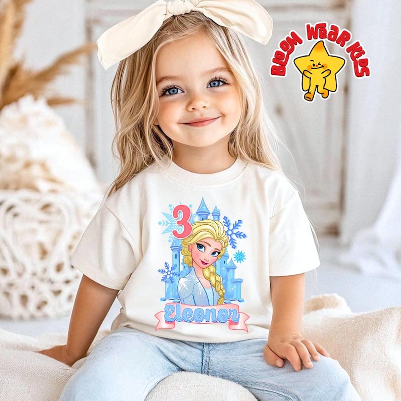 Może przedstawiać: Kremowa koszulka z nadrukiem księżniczki z kresk&oacute;wki, cyfrą 3, zamkiem i imieniem "Eleonor" na banerze. Koszulka jest noszona przez małe dziecko z blond włosami i kremową kokardą. Widoczne jest logo "BOOM WEAR KIDS".