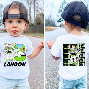 May include: White t-shirt featuring a Buzz Lightyear graphic, a large green number 3, and the name "LANDON" in black. The back of the shirt has a repeating "BIRTHDAY BOY" design with the Buzz Lightyear graphic. The child is wearing a black cap and light blue jeans.