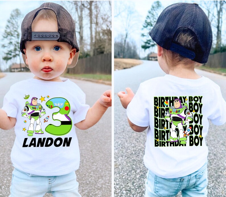May include: White t-shirt featuring a Buzz Lightyear graphic, a large green number 3, and the name "LANDON" in black. The back of the shirt has a repeating "BIRTHDAY BOY" design with the Buzz Lightyear graphic. The child is wearing a black cap and light blue jeans.