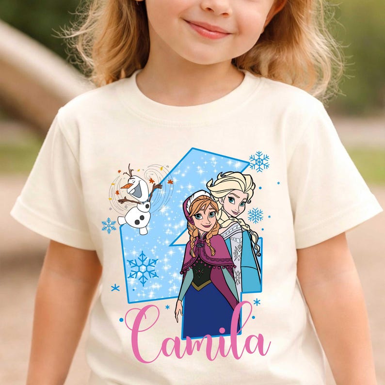 May include: A cream-colored short-sleeve t-shirt featuring a graphic of the characters Anna, Elsa, and Olaf from Frozen. The design includes a large blue number four, blue snowflakes, and the name Camila written in pink cursive script lettering.