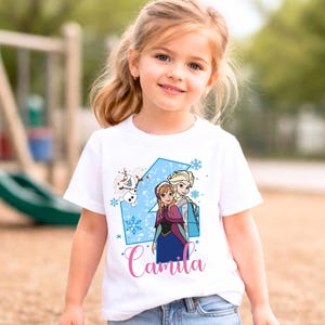 May include: A white short-sleeved t-shirt featuring a graphic print of the characters Anna, Elsa, and Olaf from Frozen. The design includes a large blue number four, light blue snowflakes, and the name Camila written in pink cursive script.