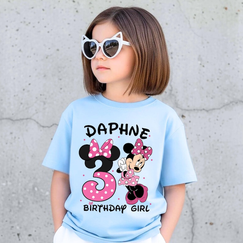 May include: A light blue t-shirt with the name "Daphne" and the number "3" in a pink polka dot design. A cartoon Minnie Mouse graphic is also on the shirt. The text "BIRTHDAY GIRL" is below the number.