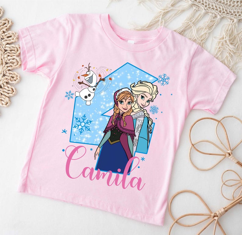 May include: A light pink short-sleeve t-shirt featuring a graphic of Anna, Elsa, and Olaf from Frozen. The design includes a large blue number three, snowflakes, and the name Camila written in a pink cursive script font across the bottom.