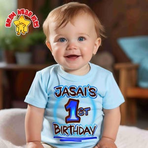 May include: A light blue t-shirt with the text "JASAI'S 1st BIRTHDAY" in brown and blue. The shirt is worn by a smiling baby with blonde hair and blue eyes. A cartoon star logo is in the upper left corner.