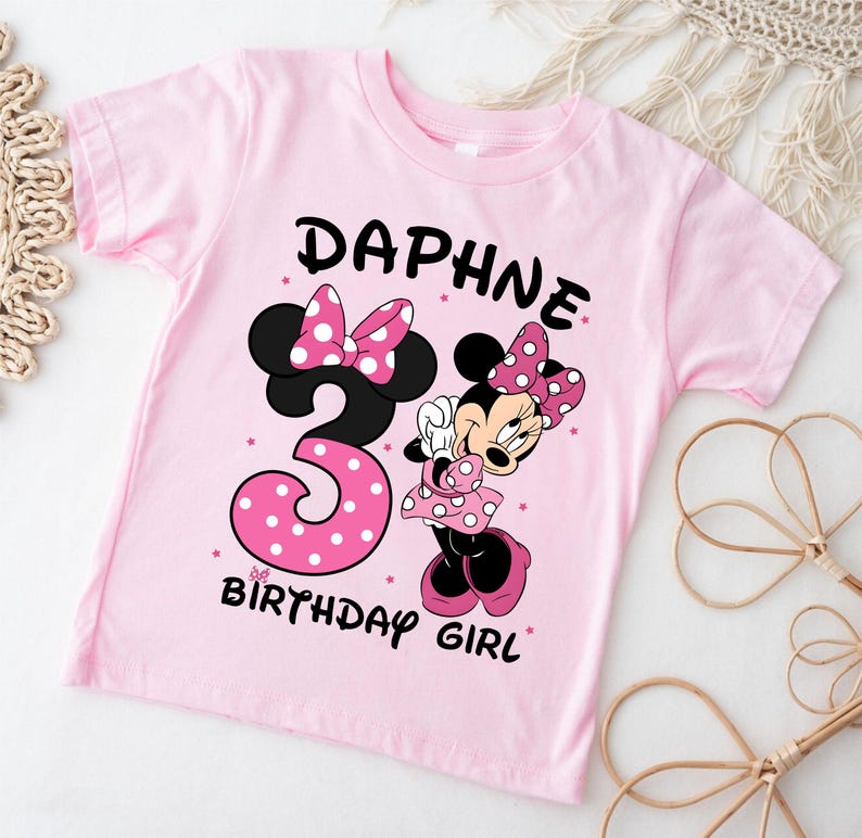 May include: A light pink t-shirt featuring a Minnie Mouse graphic. The shirt has the name "DAPHNE" above a large number "3" with pink polka dots, and the words "BIRTHDAY GIRL". Minnie Mouse is also featured on the shirt.