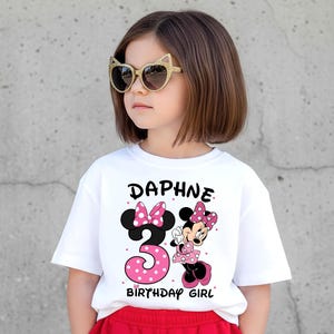 May include: A white t-shirt with the name "Daphne" and the number "3" in pink polka dots, along with a Minnie Mouse graphic. The words "BIRTHDAY GIRL" are also printed on the shirt. The child is wearing gold cat-eye sunglasses.