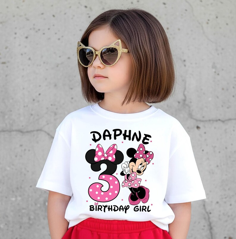 May include: A white t-shirt with the name "Daphne" and the number "3" in pink polka dots, along with a Minnie Mouse graphic. The words "BIRTHDAY GIRL" are also printed on the shirt. The child is wearing gold cat-eye sunglasses.