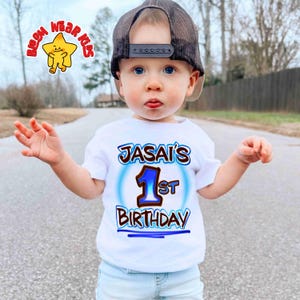 May include: A young child wearing a white t-shirt with the text "JASAI'S 1st BIRTHDAY" in blue and brown. The child is also wearing a brown mesh trucker hat. A cartoon star logo is in the upper left corner.