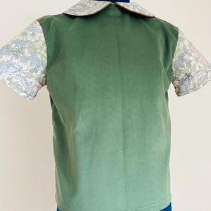 Può includere: Una camicia di velluto a coste verde con maniche a stampa floreale e colletto a stampa floreale con finiture dorate. La camicia è esposta su un manichino blu. La camicia ha maniche corte.