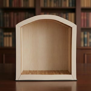 Puede incluir: Una estantería de madera de color claro con una parte superior arqueada. El interior es de un tono más claro de madera, y la estantería se encuentra sobre un fondo borroso de estanterías. La estantería está vacía.