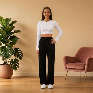 Könnte beinhalten: Eine Frau trägt ein weißes, langärmeliges Crop-Top und eine schwarze Hose mit Kordelzug. Sie trägt weiße Turnschuhe. Der Hintergrund umfasst eine große Topfpflanze und einen rosa Sessel.