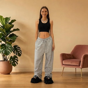 Könnte beinhalten: Eine Person trägt ein schwarzes Crop-Top, eine graue Jogginghose mit Kordelzug und schwarze, flauschige Hausschuhe mit Metallakzenten. Sie steht vor einer Topfpflanze und einem rosa Sessel.