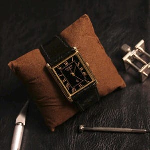 May include: A gold-toned Citizen Quartz watch with a black rectangular face and black leather band. The watch features Roman numeral hour markers. The watch is displayed on a brown pillow. Watch repair tools are also visible.